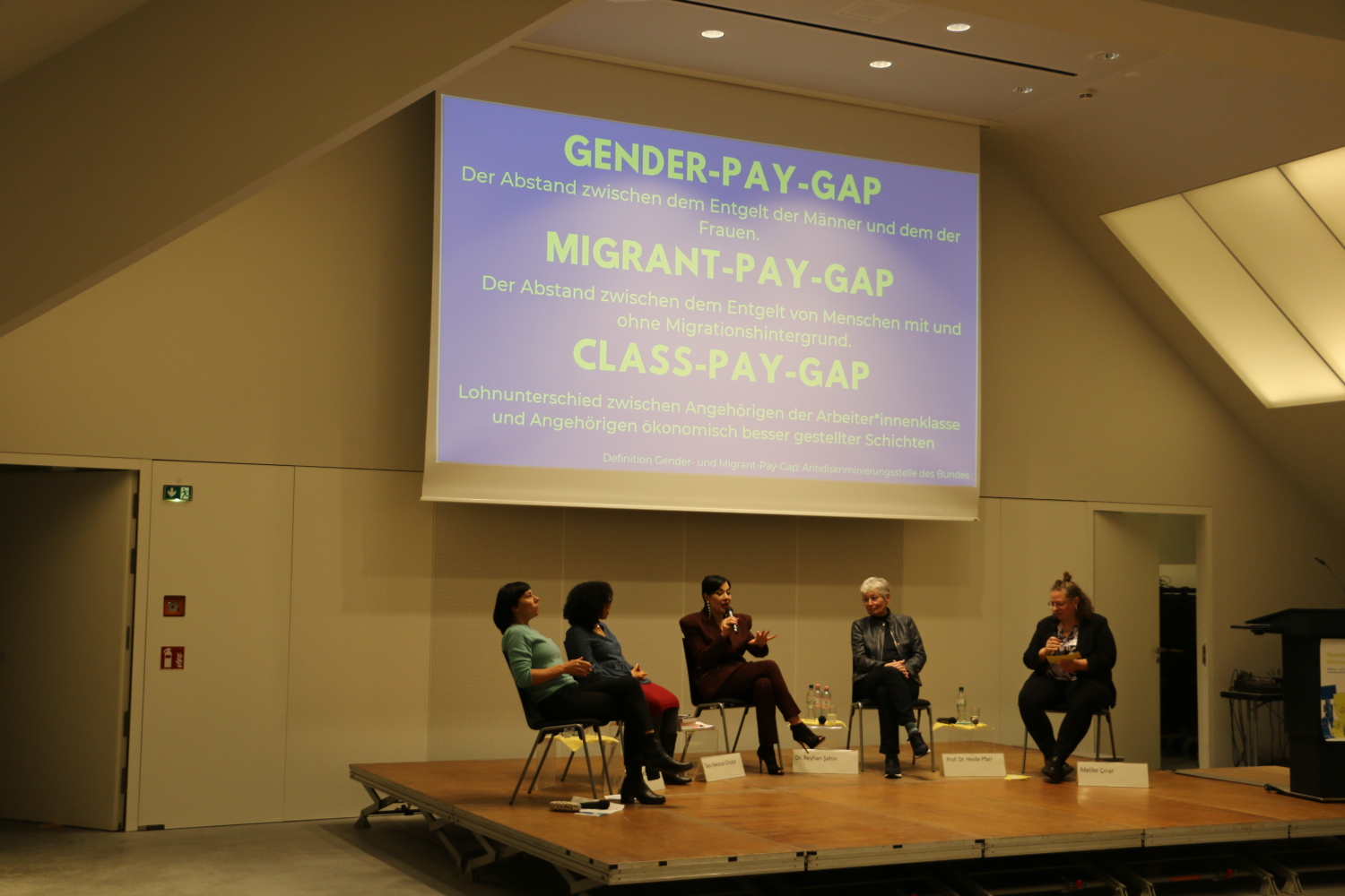 Die Podiumsgäst*innen sitzen auf der Bühne. Dr. Reyhan Şahin spricht ins Mikrofon. Im Hintergrund ist eine Präsentation auf einer Leinwand, auf der die Definitionen von Gender-, Migrant- und Class-Pay-Gap stehen.