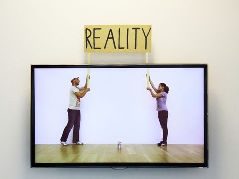 Auf einem Bildschirm heben ein Mann und eine Frau ein Banner mit dem Schriftzug "Reality" hoch, dieser Banner steigt hinter dem Bildschirm tatsächlich hoch.