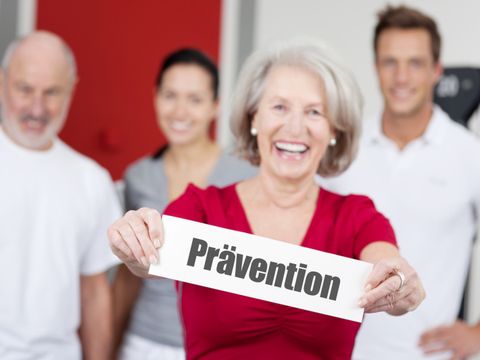 Eine Frau präsentiert ein Schild mit der Aufschrift "Prävention". Im Hintergrund stehen drei Menschen verschiedenen Alters in Sportkleidung.