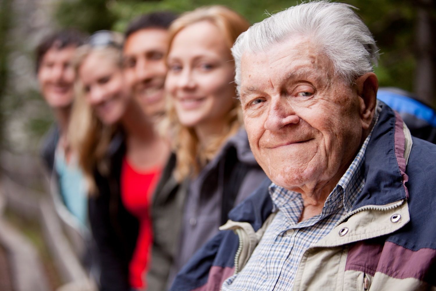 Ein älterer Mann sitzt zusammen mit einer Gruppe jüngerer Menschen und alle lachen Richtung Kamera