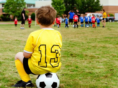 Ein kleiner Junge, auf einem Fußball sitzend, schaut zu einer Gruppe von Fußballern 