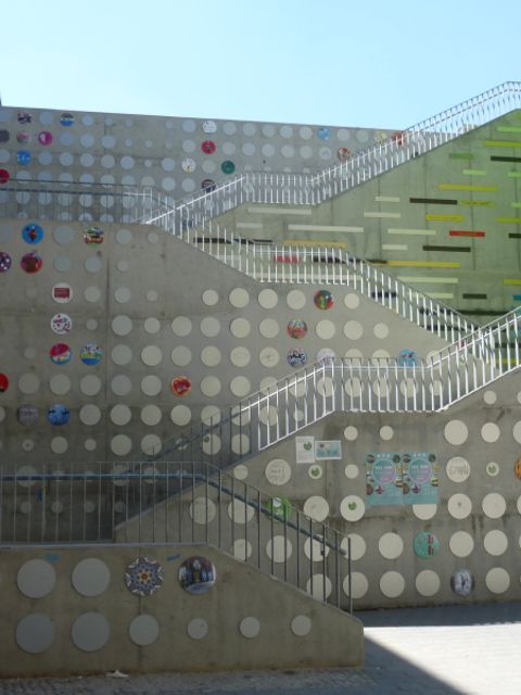 Bildvergrößerung: Treppe zwischen einer Bachsteinwand und einer grün angemalten Wand. Die Treppe führt über vier Treppenläufe nach oben. An der Rückseite der Treppe sind runde Scheiben mit Illustrationen von Kindern und Jugendlichen anmontiert.