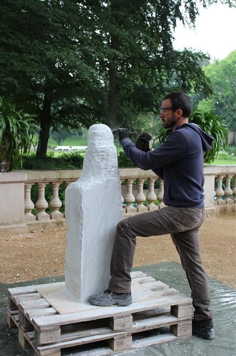 Bildhauer Daniel Bayardi bearbeitet stehend eine Steinskulptur, im Hintergrund die grünen Bäume des Körnerparkes