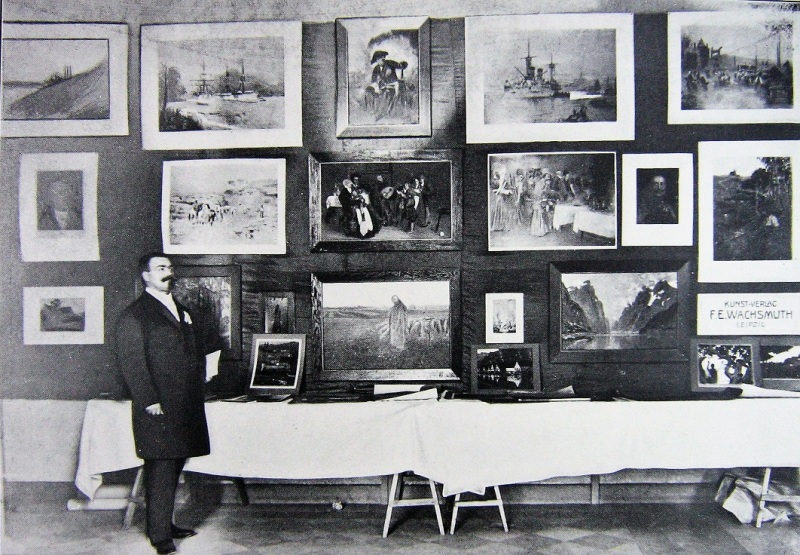 Der Museumsleiter Emil Fischer steht vor einer Wand, an der Gemälde hängen, neben sich einen Tisch, auf dem Dokumente präsentiert werden