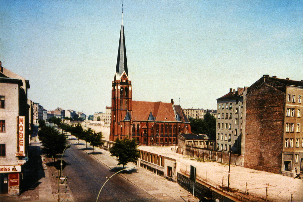 Bernauer Straße in Höhe der Ackerstraße um 1970: Bis auf die Versöhnungskirche wurde ab 1965 die Bebauung auf dem Grenzstreifen abgerissen und nur Teile der Straßenfassaden als vorläufige Mauer stehen gelassen.