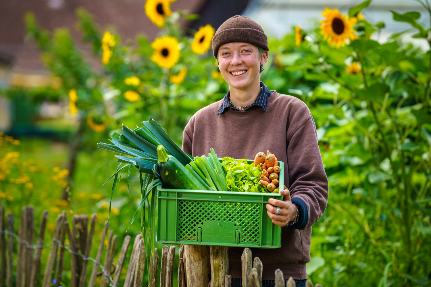 Eine Frau trägt eine Kiste mit frisch geertnetem Gemüse, im Hintergrund Sonnenblumen
