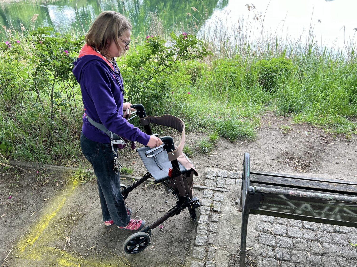 Der Boden um eine Sitzbank am Orankesee ist uneben und dicht bewachsen. Dies erschwert die Zugänglichkeit für Rollatornutzer:innen. Die Barriere wurde mit gelbem Kreidespray kenntlich gemacht.