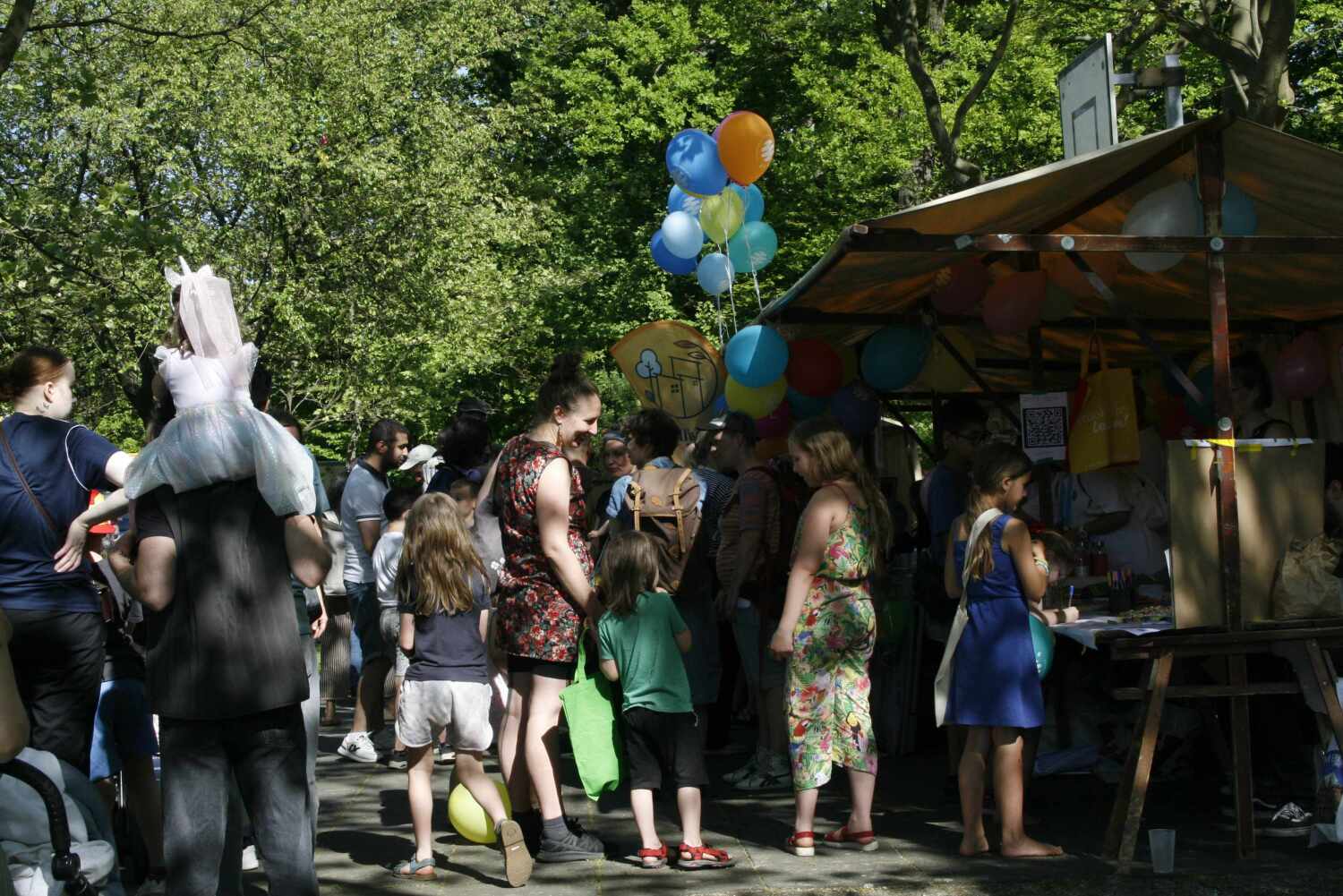 Maifest 2024 - Menschen stehen am Stand an, Frau mit Kindern im Vordergrund, am Stand hängen Luftballons
