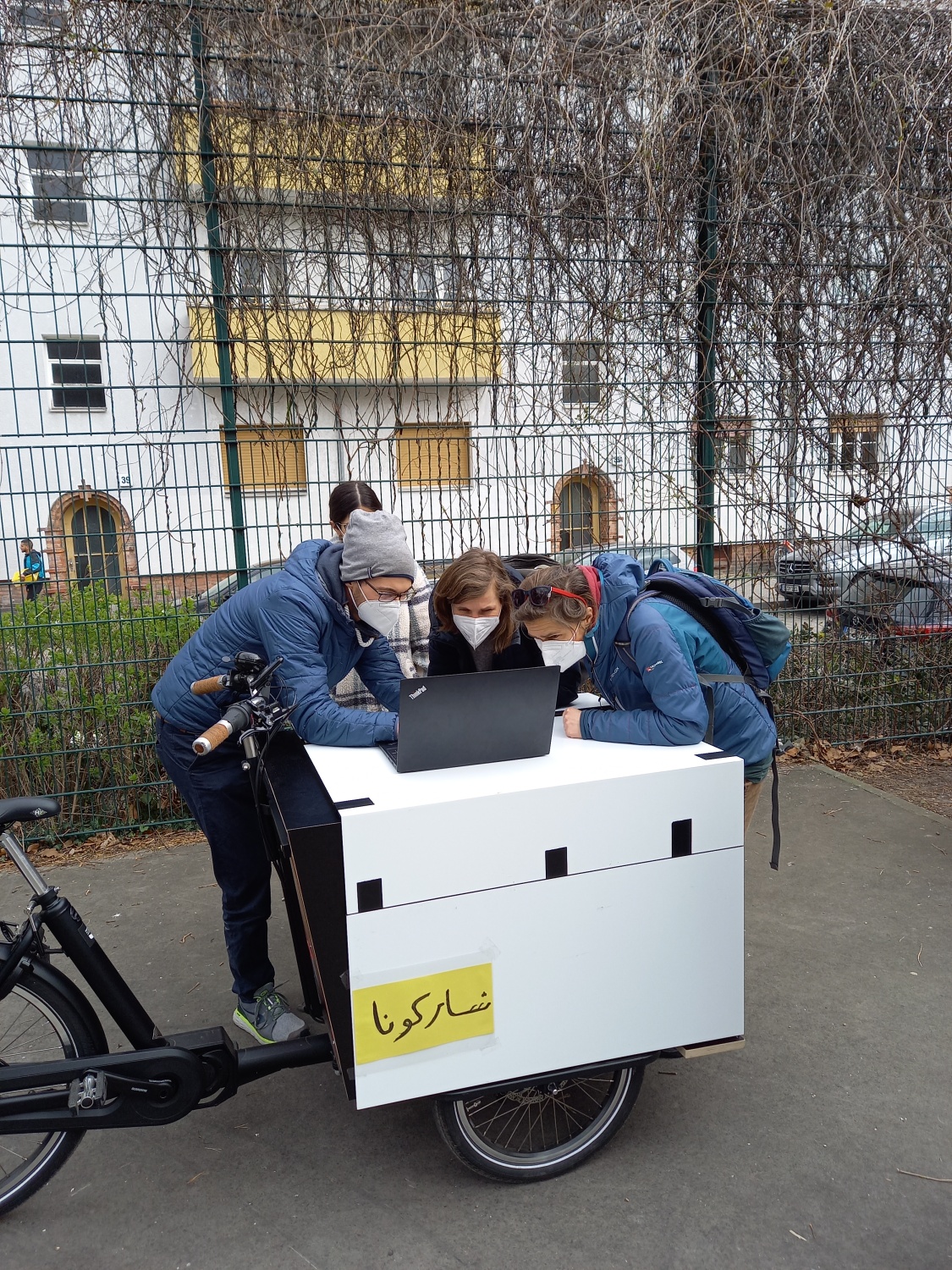 Vier Personen schauen auf einen Laptop der auf einem Lastenrad auf einem Sportplatz steht. 