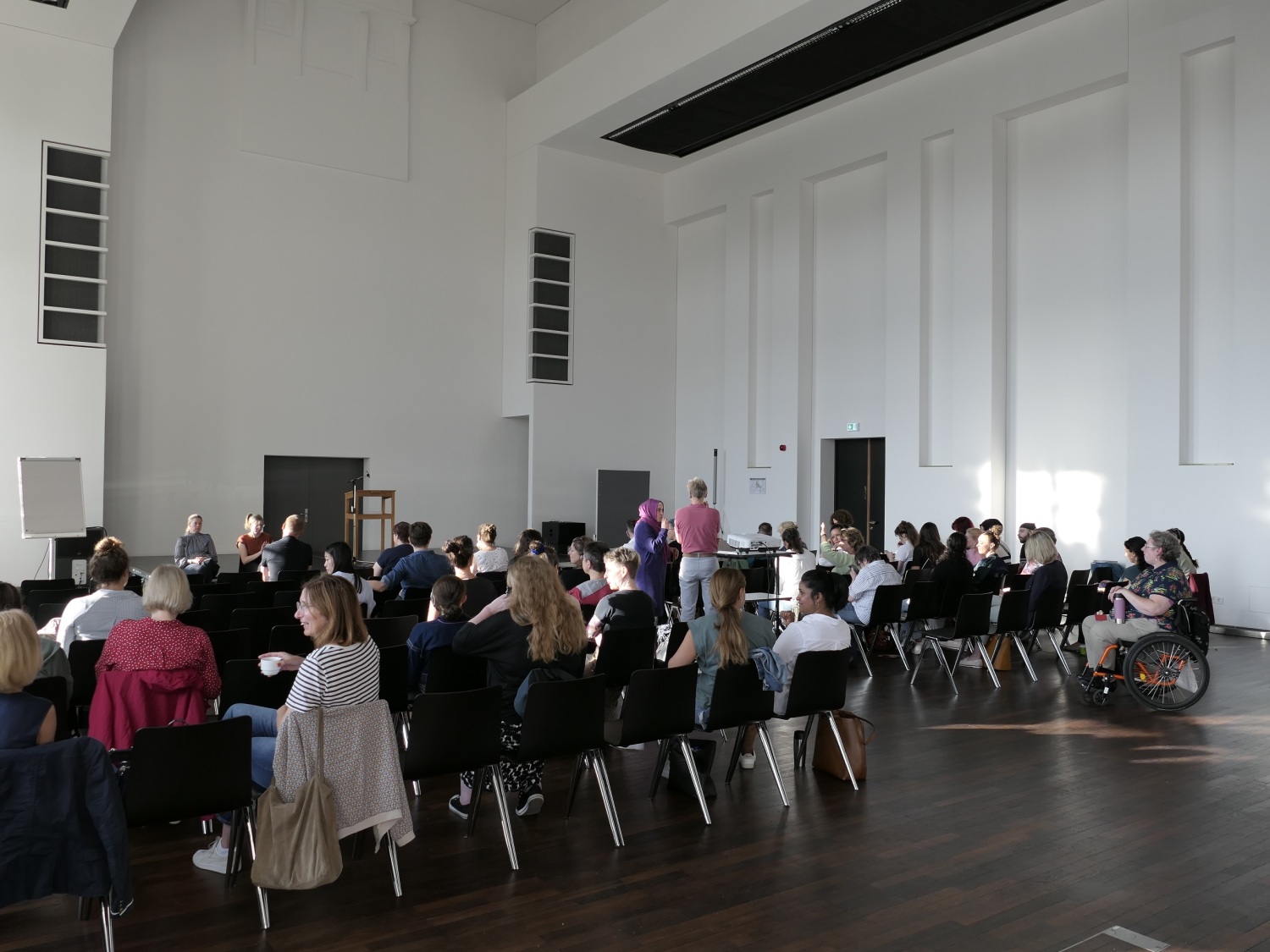 In einem großen Saal sind mehrere Stuhlreihen vor einer Bühne. Die Veranstaltung ist gut besucht.