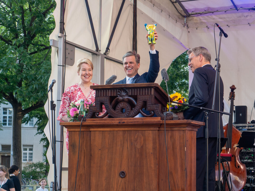 Drei Menschen stehen auf einer Bühne hinter einem hölzernen Rednerpult. Der Mann in der Mitte hält stolz eine kleine Bären-Skulptur in die Höhe.