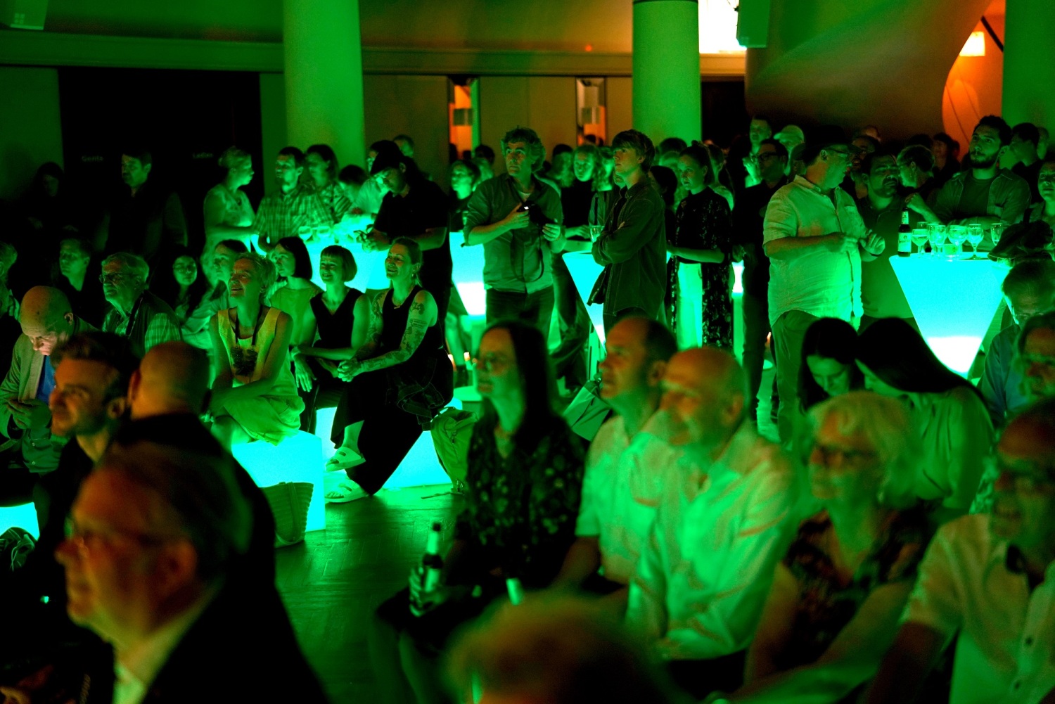In einem dunklen grün beleuchteten Saal sitzen und stehen Menschen und schauen in eine Richtung.