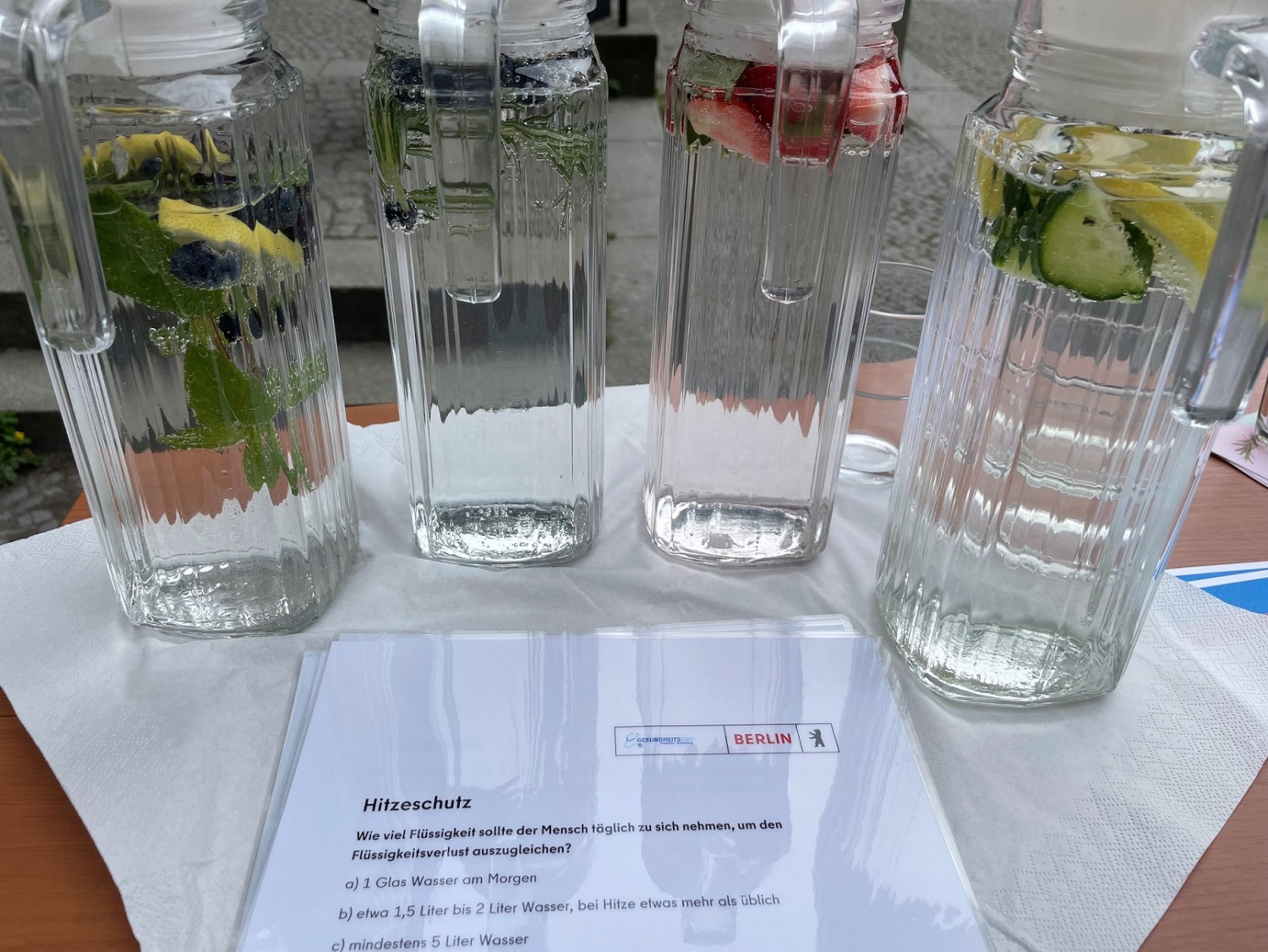 Auf einem kleinen Stand stehen vier Glaskaraffen mit Wasser und Früchten und ein laminiertes Quiz zum Thema Hitzeschutz.