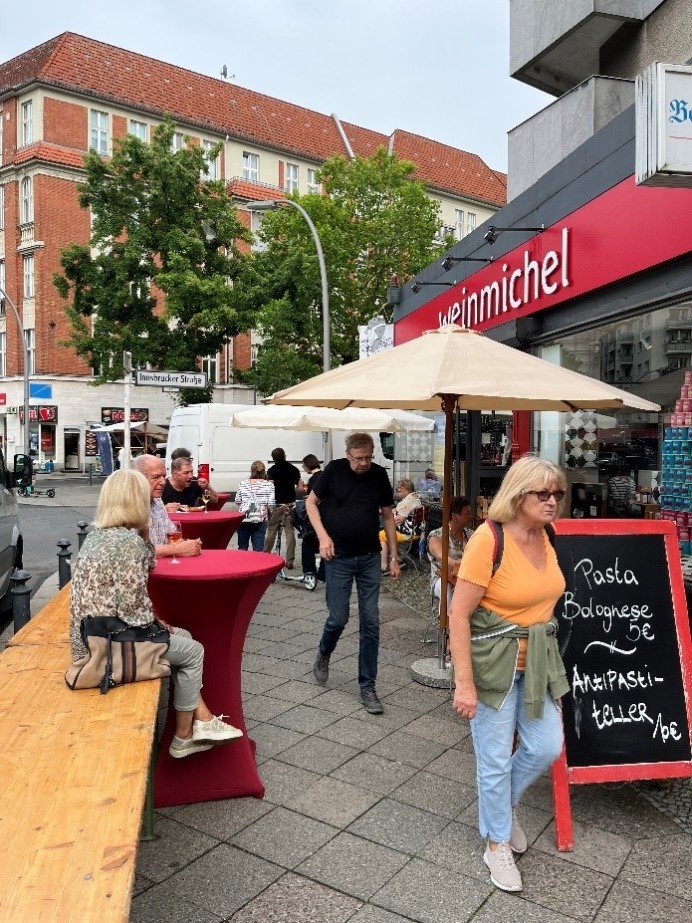 Vor einem Lokal sitzen mehrere Personen draußen auf Stühlen und an Stehtischen und genießen Wein, Snackplatten und vieles mehr.