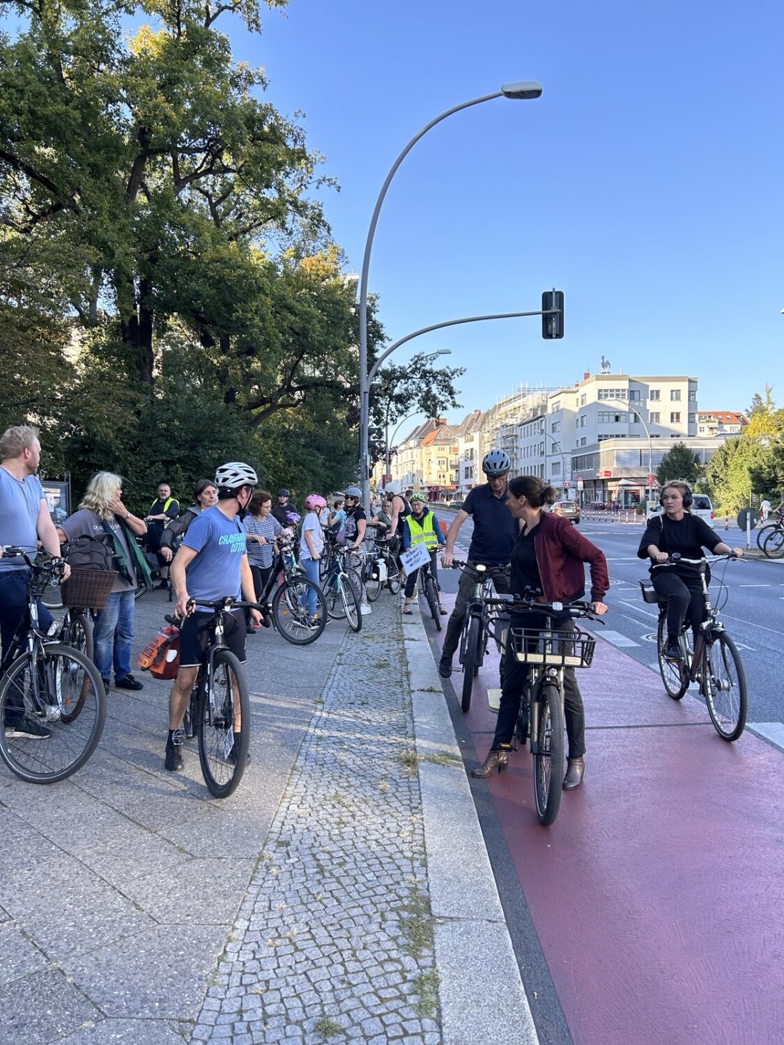 Viele Personen auf Fahrrädern stehen auf einem Gehweg neben einem Radweg.