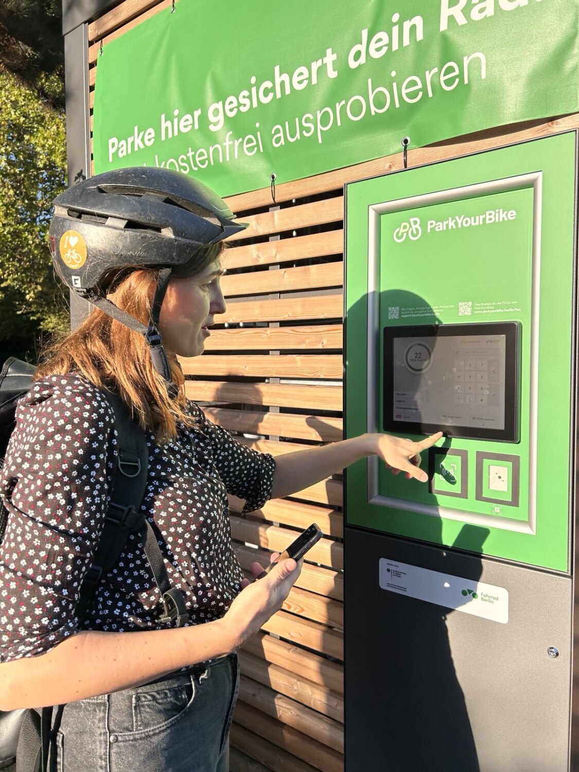 Eine Frau mit Fahrradhelm steht vor einem Container und schaut auf ein Bedienpult. Sie hält ein Handy in einer Hand und tippt mit der anderen etwas auf einem Touchscreen an.