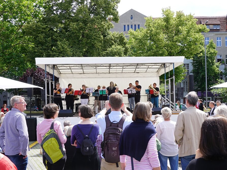 viele Menschen stehen vor einer Open-Air-Bühne, auf der veile Personen mit Blasinstrumenten stehen.