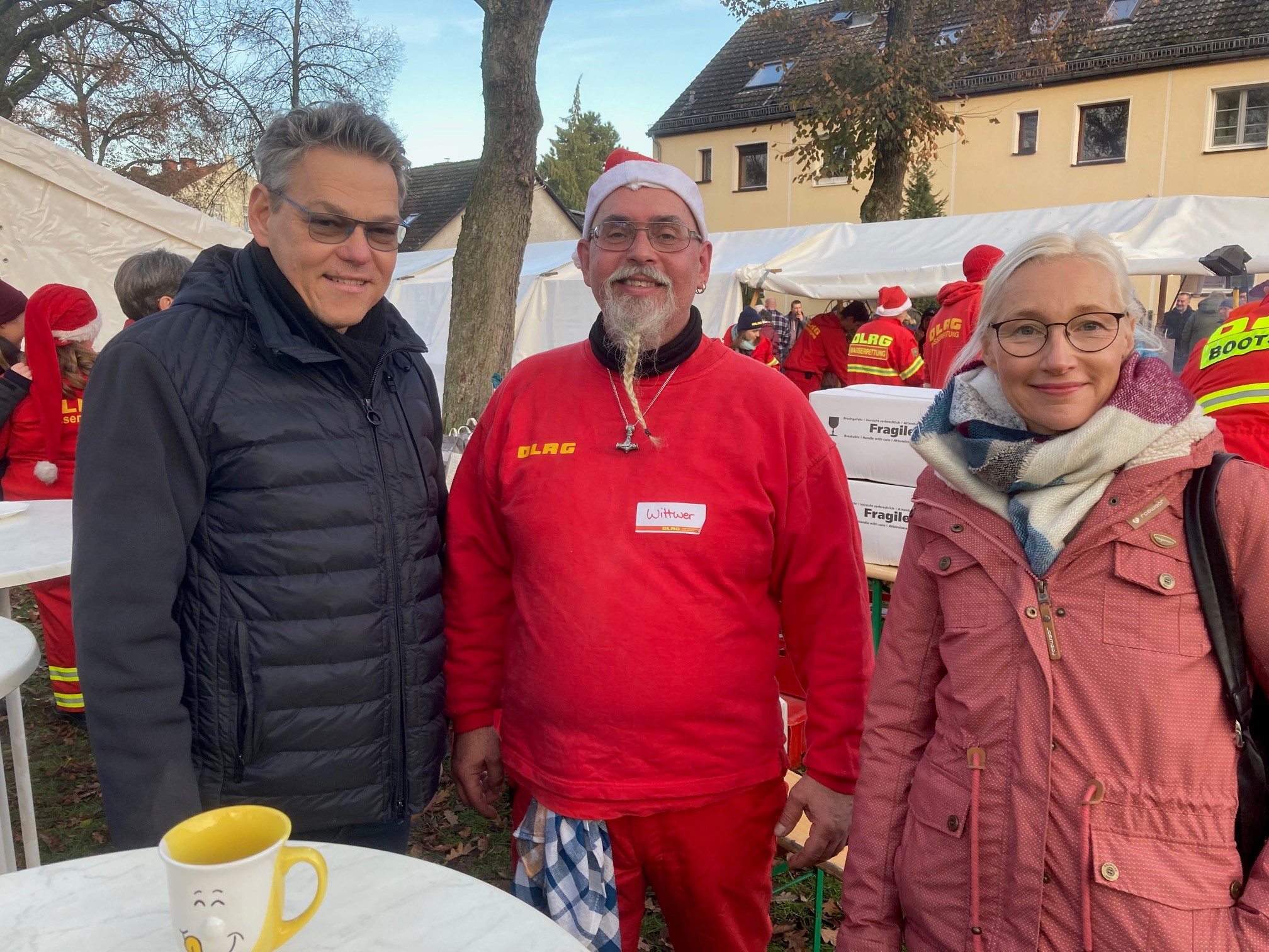 Mann in rotem Pulli und mit Nikolausmütze steht in der Mitte zwischen einem Mann und einer Frau