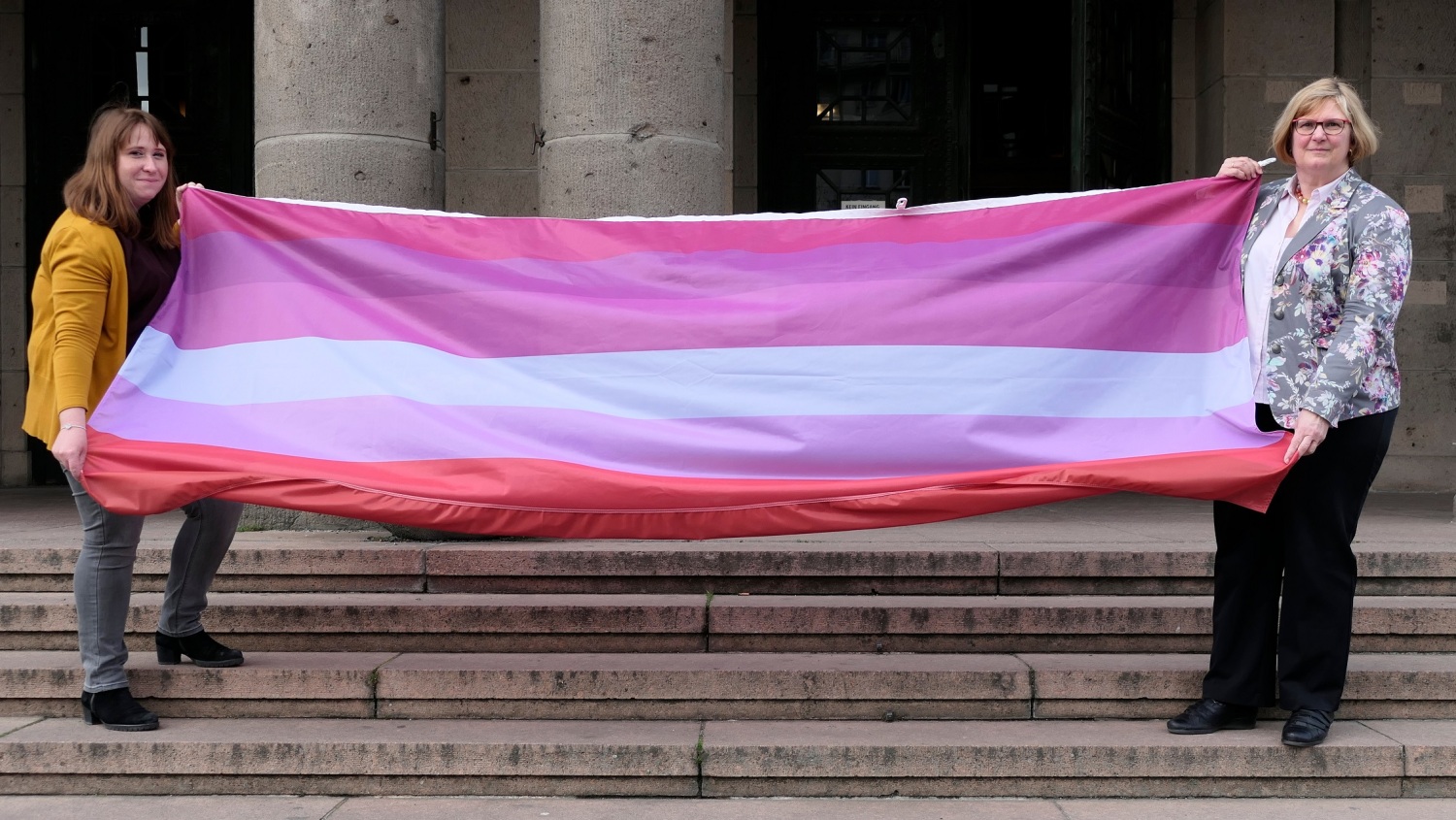 Zwei Frauen stehen auf einer Treppe vor einem Gebäude und halten zusammen eine rot-orange-pink-weiß gestreifte Flagge hoch.