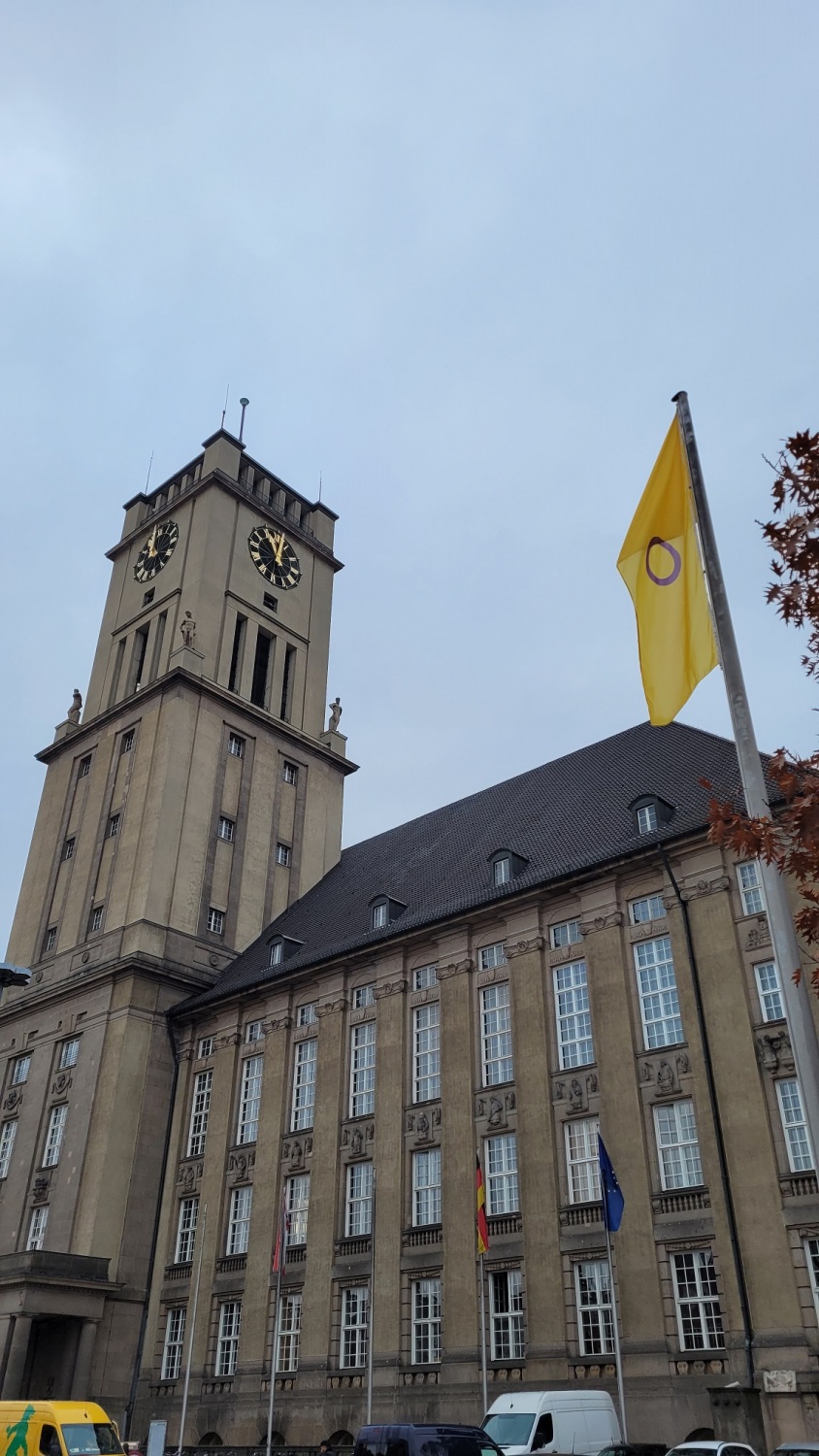 Vor einem großen Gebäude mit Uhrenturm weht eine gelbe Flagge mit lila Kreis an einem Fahnenmast.