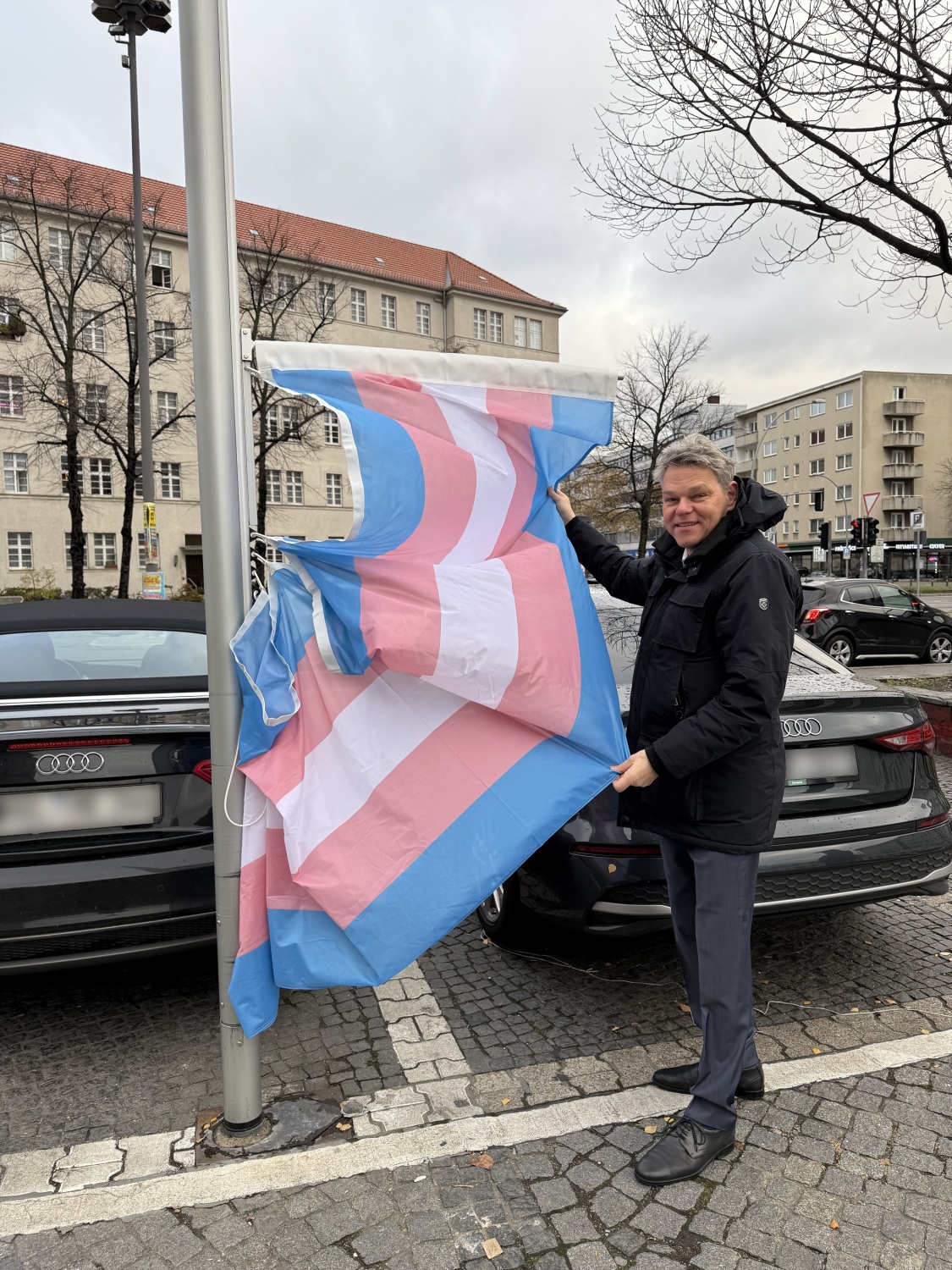 Ein Mann steht neben einem Fahnenmast und hält daran eine blau-rosa-weiß gestreifte Flagge hoch.