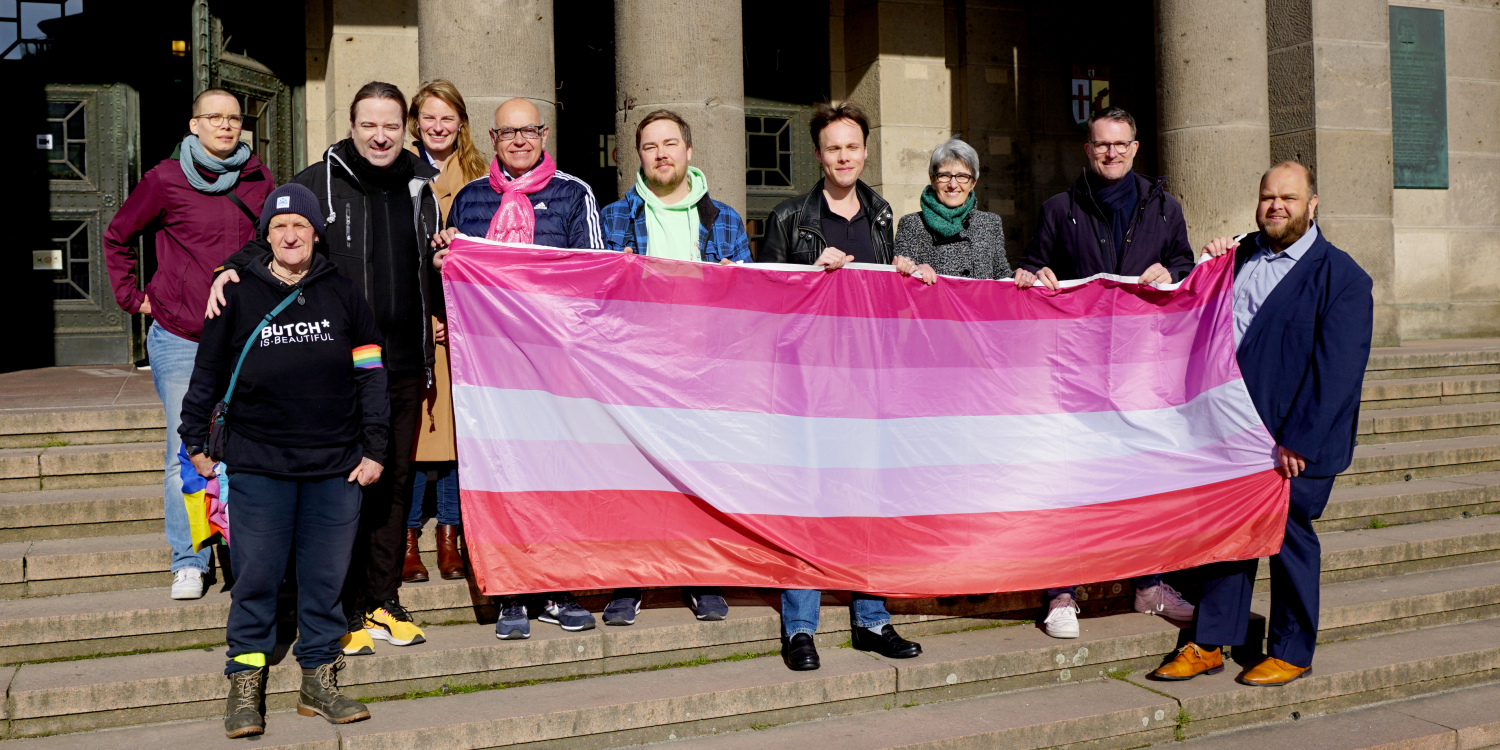 Eine Gruppe Menschen steht auf der Vortreppe eines Gebäudes. Zusammen wird eine Flagge mit pinken, orangenen und einem weißen Streifen in der Mitte hochgehalten.