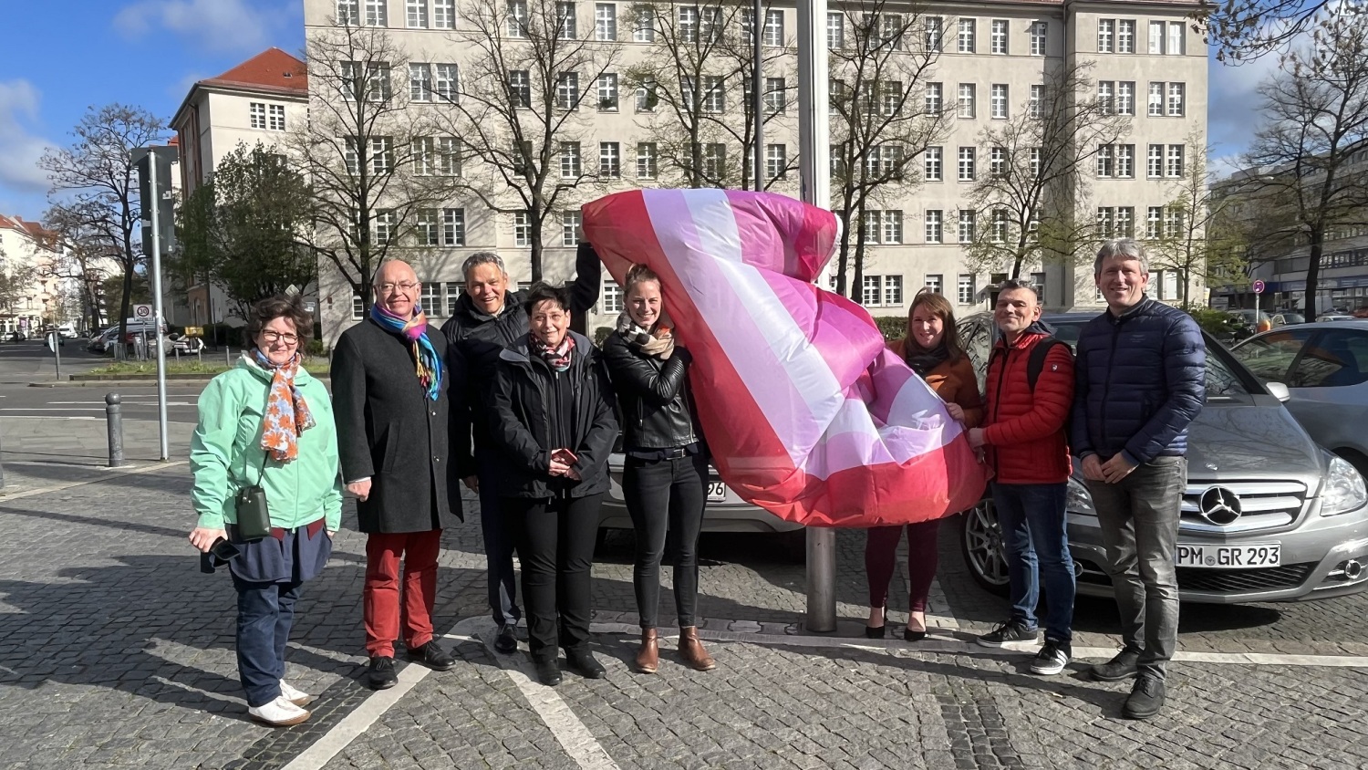 Eine Gruppe Menschen steht mit einer rot-orange-pink-weißen Fahne vor einem Fahnenmast.