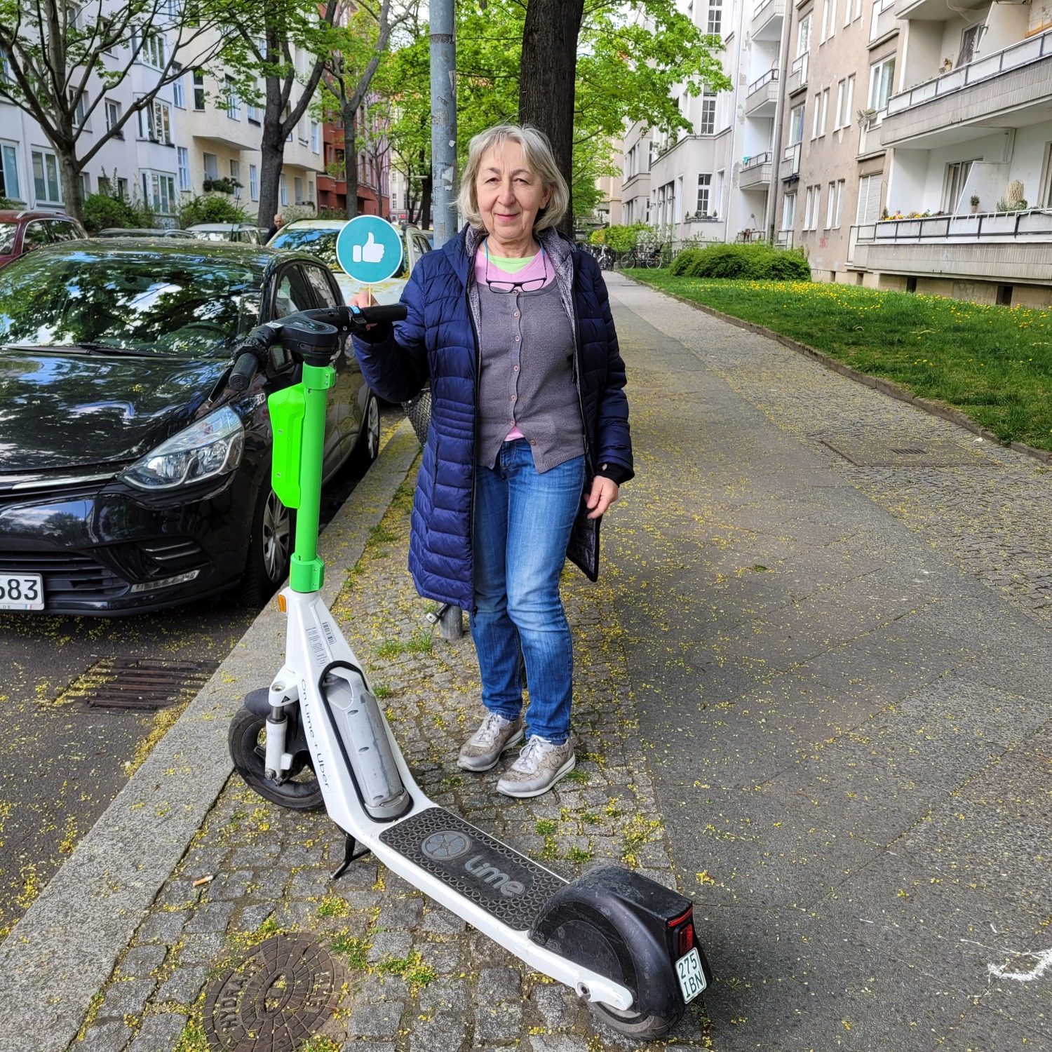 Eine Frau mit grauen Haaren, blauer Jacke und Jeans steht auf dem Gehweg. Sie hat ein rundes grünes Schild in der rechten Hand. Darauf ist ein Daumen, der nach oben zeigt, zu sehen. Vor ihr steht ein grün weißer E-Roller. Der Gehweg ist frei.