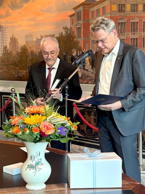 Zwei Männer stehen vor einem farbigen Wandgemälde. Der eine hält eine geöffneteSchatulle mit einem Orden, der andere hält eine Mappe und spricht in ein Mikrofon. Vor ihnen auf dem Tisch ein bunter Blumenstrauß und ein Geschenk.