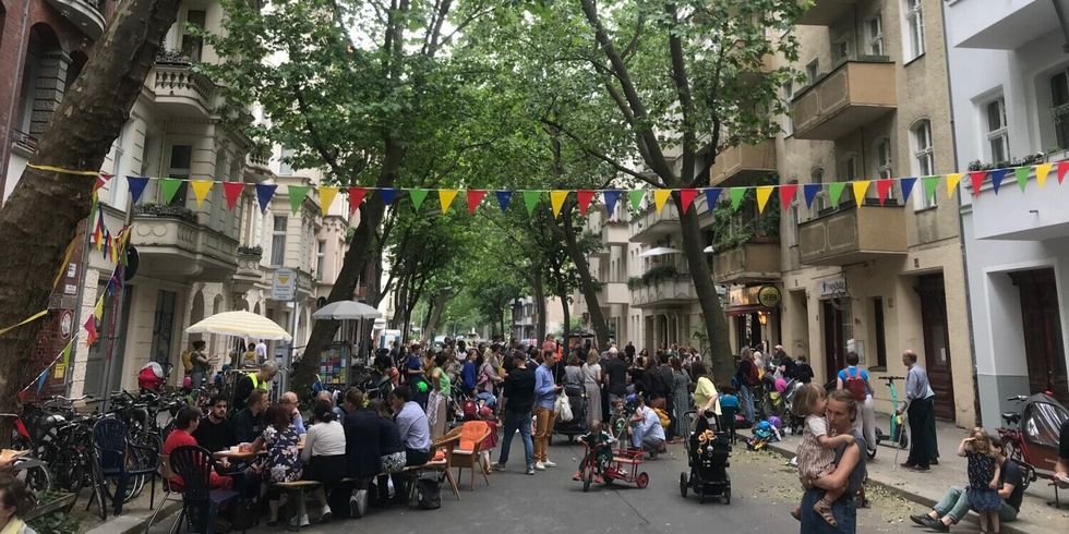 Auf einer Straße laufen und sitzen viele Menschen zusammen und spielen und unterhalten sich. Die Straße ist mit Wimpeln geschmückt.