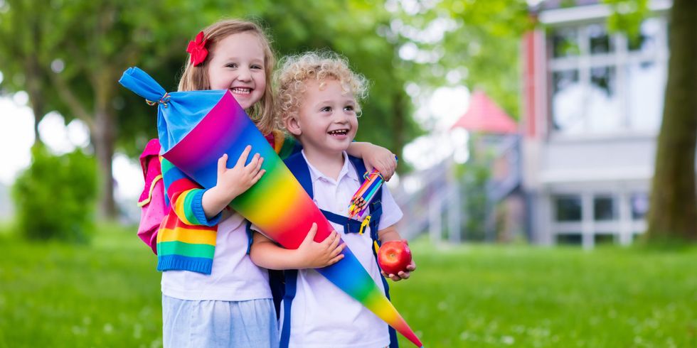 Zwei lachende Kinder mit einer regenbogenfarbenen Schultüte im Arm auf einer Wiese