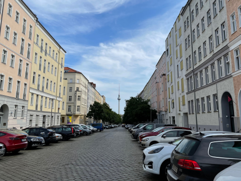 eine leere Straße mit Häusern und parkenden Autos an den Seiten. Im Hintergrund ist in der Mitte der Straße der Fernsehturm zu sehen.