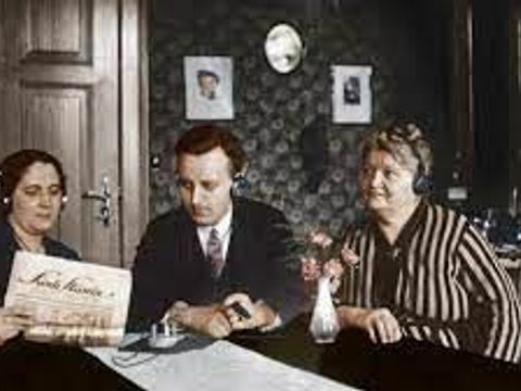  Das zeitgenössische nachkolorierte Foto aus den 20er Jahren des 20.Jhd. zeigt zwei Frauen und einen Mann an einem Wohnzimmertisch mit Kopfhörern sitzen, die der Funkstunde-Berlin lauschen, deren Programmzeitschrift eine der Frauen gut sichtbar in Händen hält.