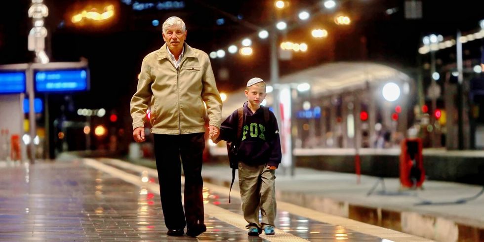 Ein älterer Mann und ein kleiner Junge laufen Hand in Hand auf einem Bahnhof an einem Gleis entlang.