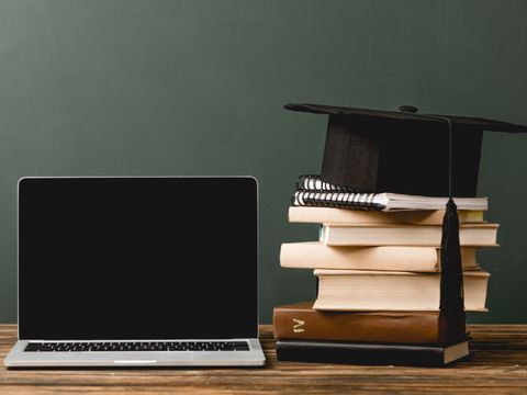 Laptop und Bücherstapel auf einem Tisch, akademische Mütze liegt auf dem Bücherstapel