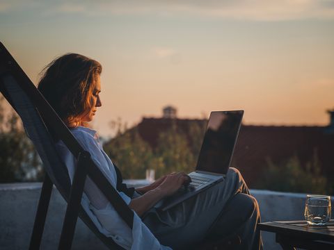 Frau auf Dach in Liegestuhl mit Laptop, Sonnenuntergang