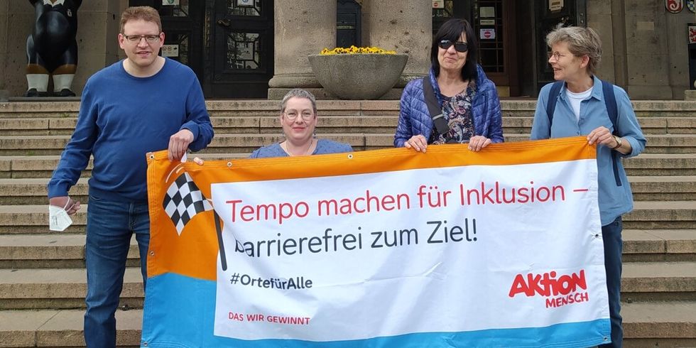 Vier Personen halten ein Banner auf dem steht: „Tempo machen für Inklusion – barrierefrei zum Ziel. #OrteFürAlle Das Wir gewinnt.“ Sie stehen vor den Treppen des Rathauses Schöneberg. Eine Person sitzt im Rollstuh. Alle lachen in die Kamera.