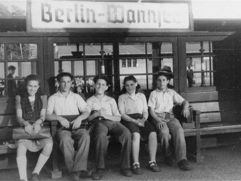 Bildvergrößerung: Zwei junge Frauen und drei junge Männer sitzen auf einer Holzbank vor einem Gebäude mit dem Schild "Berlin-Wannsee".