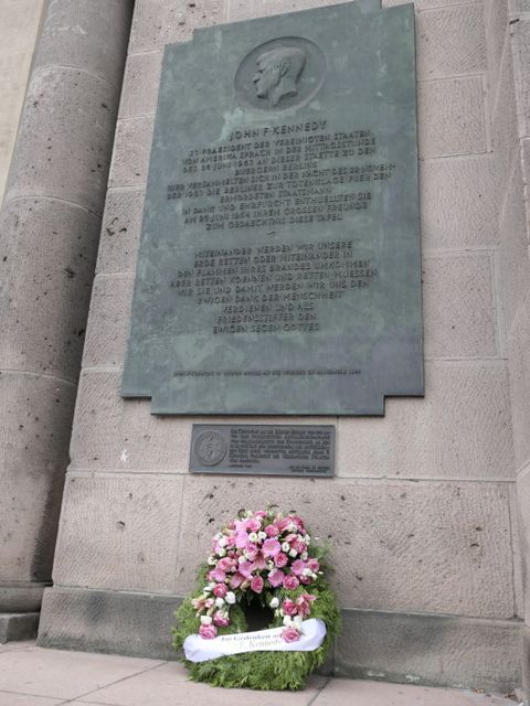 Bildvergrößerung: Ein Kranz mit Blumen lehnt an einer Wand unter einer Gedenktafel zu John-F.-Kennedy.