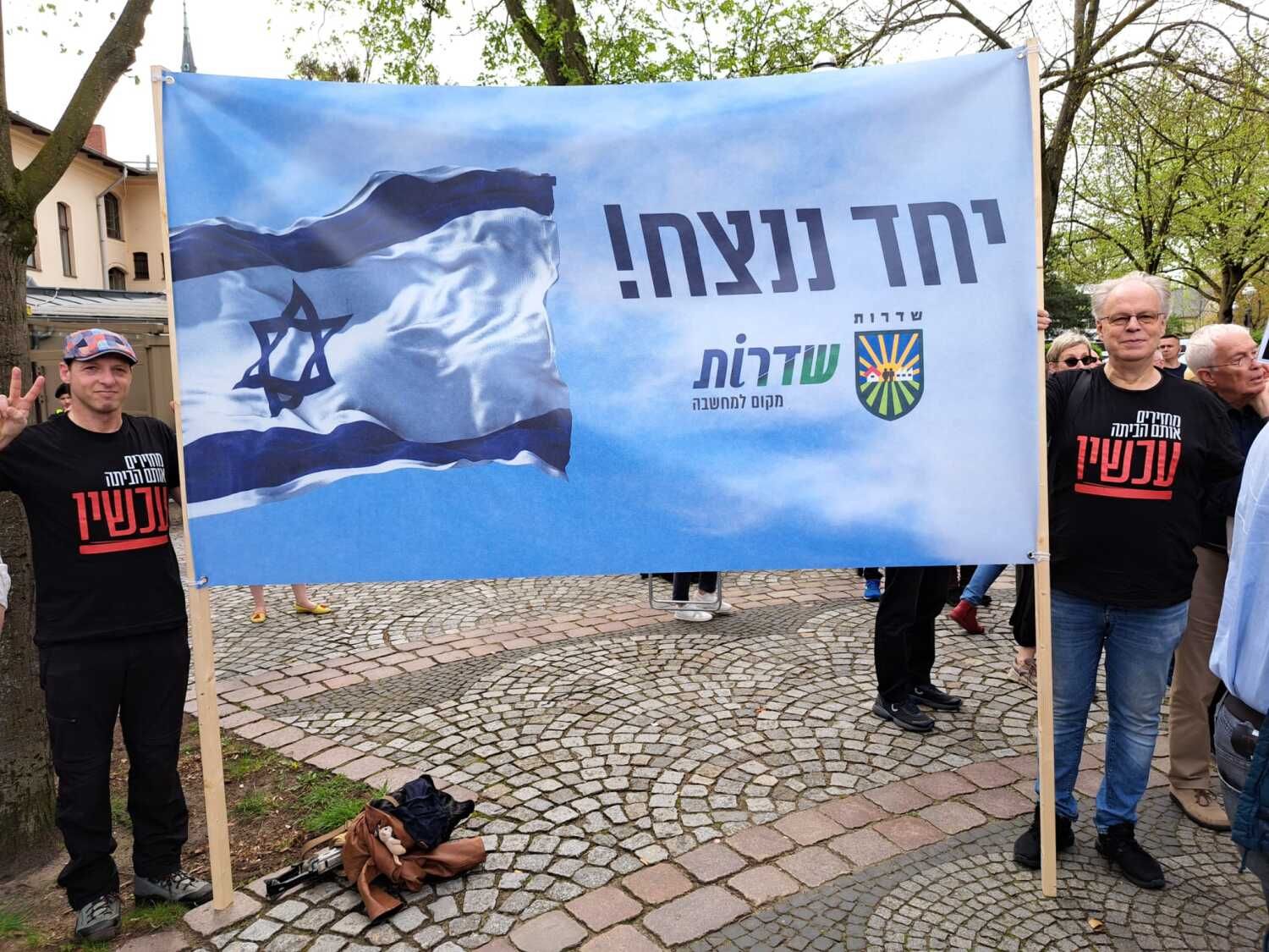 7. April 2024 Solidaritätskundgebung auf dem Sderotplatz: Jan Kutscher (Deutsch-Israelische Gesellschaft Berlin und Brandenburg e.V., auf dem Bild rechts) mit einem Transparent, auf dem das Wappen von Sderot und ein programmatischer Leitspruch aufgedruckt sind