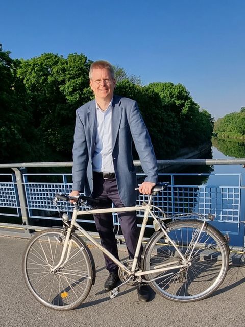 Bildvergrößerung: Ein Mann steht neben einem Fahrrad auf einer Brücke. Er lächelt in die Kamera.