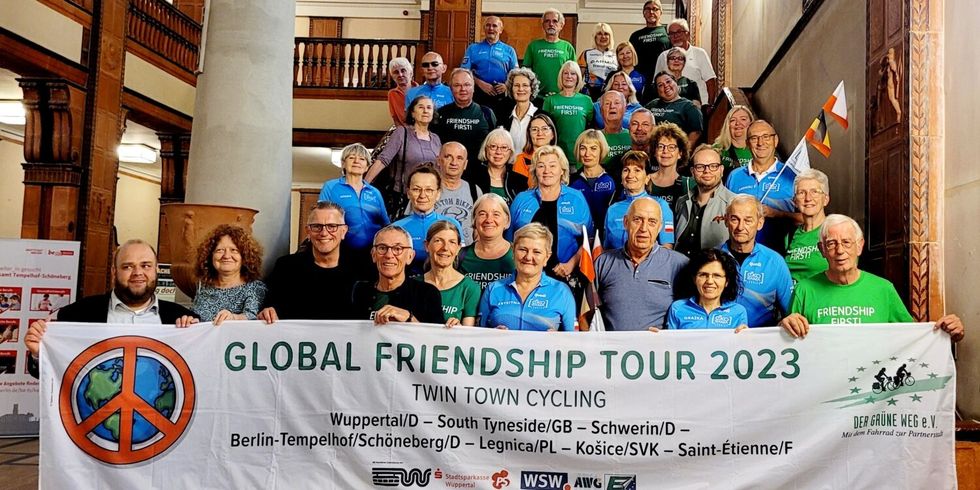 Eine Menschengruppe steht in einem großen Foyer auf einer Treppe. Im Vordergrund wird ein Banner hochgehalten, auf dem "Global Friendship Tour 2023" steht.