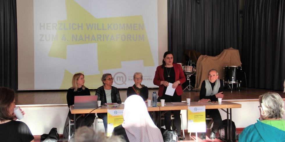 An einer Tischreihe vor einer Bühne sitzen vier Personen. Eine Frau in einem roten Jackett steht und redet. Im Hintergrund steht an einer Leinwand "Willkommen zum 4. Nahariyaforum".