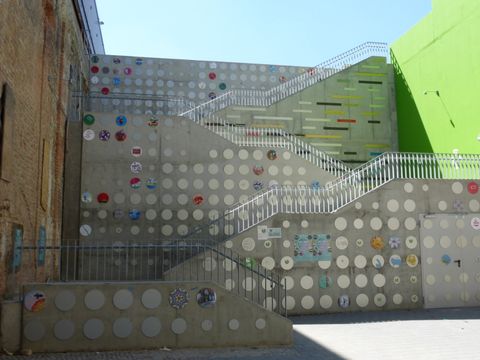 Treppe zwischen einer Bachsteinwand und einer grün angemalten Wand. Die Treppe führt über vier Treppenläufe nach oben. An der Rückseite der Treppe sind runde Scheiben mit Illustrationen von Kindern und Jugendlichen anmontiert.