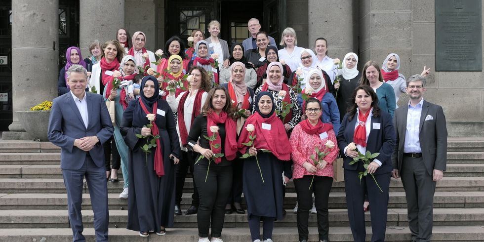 Eine Gruppe von Menschen steht auf den Treppen vor einem Gebäude und schauen in die Kamera. Viele Frauen tragen einen roten Schal und haben eine Blume in der Hand.