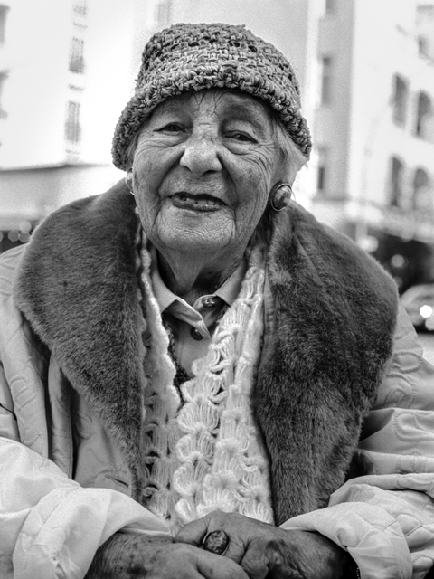 Bildvergrößerung: Eine schwarz-weiß-Fotografie von einer alten Frau mit Mütze und dicker Jacke.