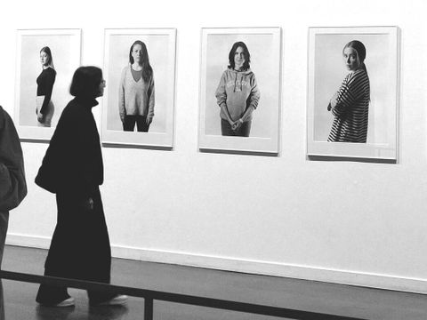 Bildvergrößerung: Schwarzweißfotografie von einer Frau, die auf eine Wand schaut. Dort sind vier Bilderrahmen mit Fotos von Frauen zu sehen.