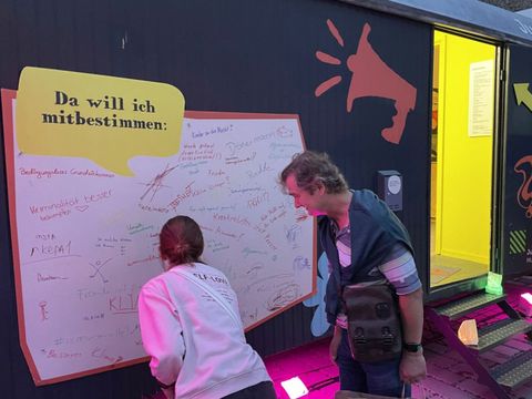 Bildvergrößerung: Zwei Menschen stehen neben einem Container und schauen auf eine weiße Fläche unter der Überschrift "Da will ich mitbestimmen:". Auf der Fläche stehen viele verschiedenen Sachen geschrieben.