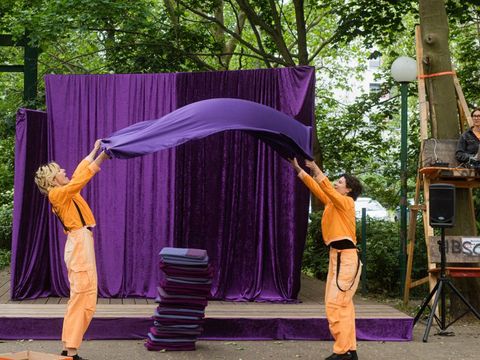Zwei Künstlerinnen in orangenen Overalls schwingen ein lila Tuch vor einer Bühne im Freien.