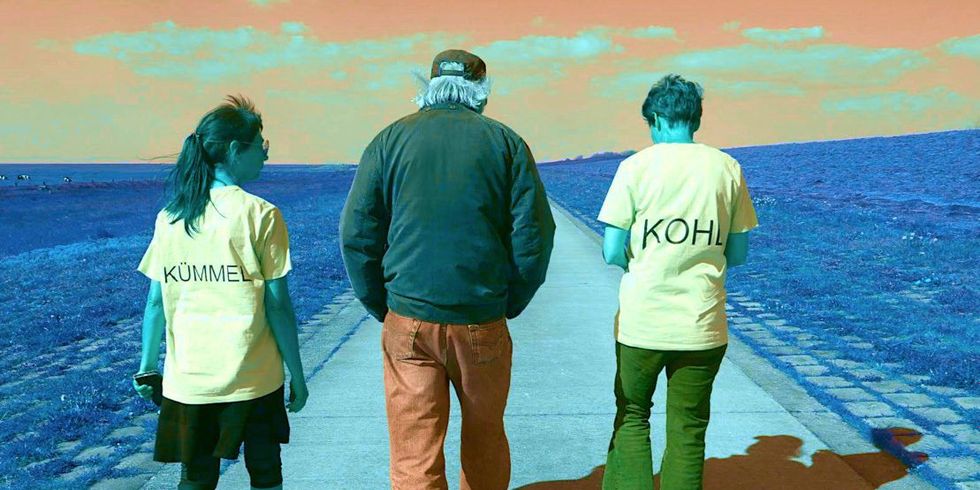 Das Bild zeigt drei Menschen von hinten, die in ein Gespräch vertieft auf einem Weg in Richtung Horizont laufen. Die beiden äußeren Personen tragen je ein gelbes T-Shirt mit den Aufschriften "Kümmel" und "Kohl".